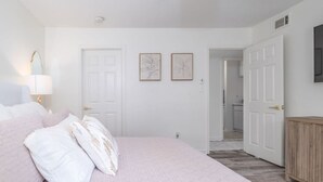 Ein Schlafzimmer mit einem Bett, einem Nachttisch, zwei gerahmten Bildern und einer Tür zu einem Flur.