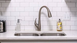 Eine Küche mit weiß gefliestem Spritzschutz, einem Edelstahl-Wasserhahn, einem gerahmten Bild über der Spüle und einer Handseifenflasche auf der Arbeitsfläche.