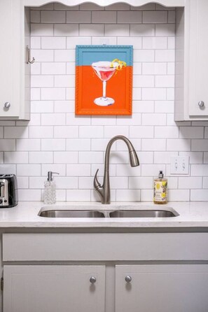A kitchen with a white tiled backsplash, a stainless steel faucet, a framed picture above the sink, and a bottle of hand soap on the counter. - Luxury in the Heart of Homewood! (Birmingham)