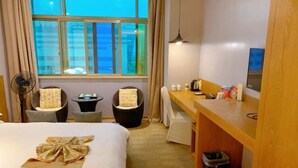 Ein Hotelzimmer mit Bett, Schreibtisch, Stuhl, einem kleinen Tisch, einem Ventilator, einem Fenster mit Vorhängen und einem Papierkorb.