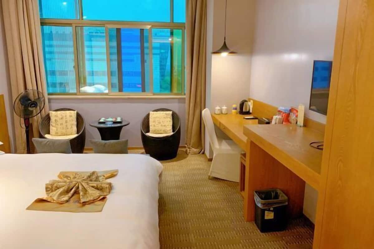Une chambre d’hôtel comprenant un lit, un bureau, une chaise, une petite table, un ventilateur, une fenêtre avec des rideaux et une poubelle.
