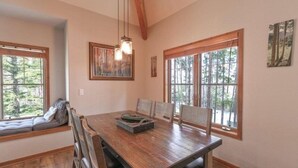 A dining area with a wooden table, chairs, and a bench with cushions. There are large windows with a view of trees. - Premier 2 Bedroom Townhome Across From Snowflake Lift - Walk to Lifts (Breckenridge)