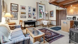 A cozy living room with a sofa, coffee table, and a dining area with chairs and a table. There is a fireplace, a kitchen area with a stove, and a staircase. - Opera House Loft 02 - Bethel Village (Bethel)