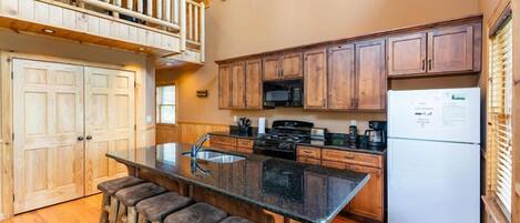 A kitchen with wooden cabinets, a black countertop, a stainless steel sink, and a refrigerator. There are wooden stools at the counter.