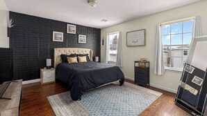 Un dormitorio con pared de ladrillo oscuro, cama con cabecera capitonada, mesita de noche, cómoda, ventana con cortinas y vistas al exterior.