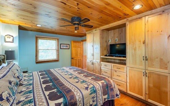 A bedroom with a bed, a ceiling fan, a TV, wooden cabinets, and a window with blinds. - Deluxe Lakefront Cabin (Vonore)