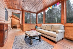 A log cabin interior with a sofa, coffee table, and large windows. - Outdoor Pool | Pool Table | Firepit | Screened Porch (Bethel)