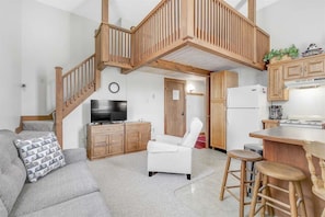 A living room with a sofa, a TV, wooden cabinets, a kitchen area with a fridge, and a staircase. - Opera House Loft 03 - Bethel Village (Bethel)
