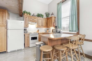 A kitchen with wooden cabinets, a white refrigerator, a stove, a microwave, and a bar area with stools. - Opera House Loft 03 - Bethel Village (Bethel)