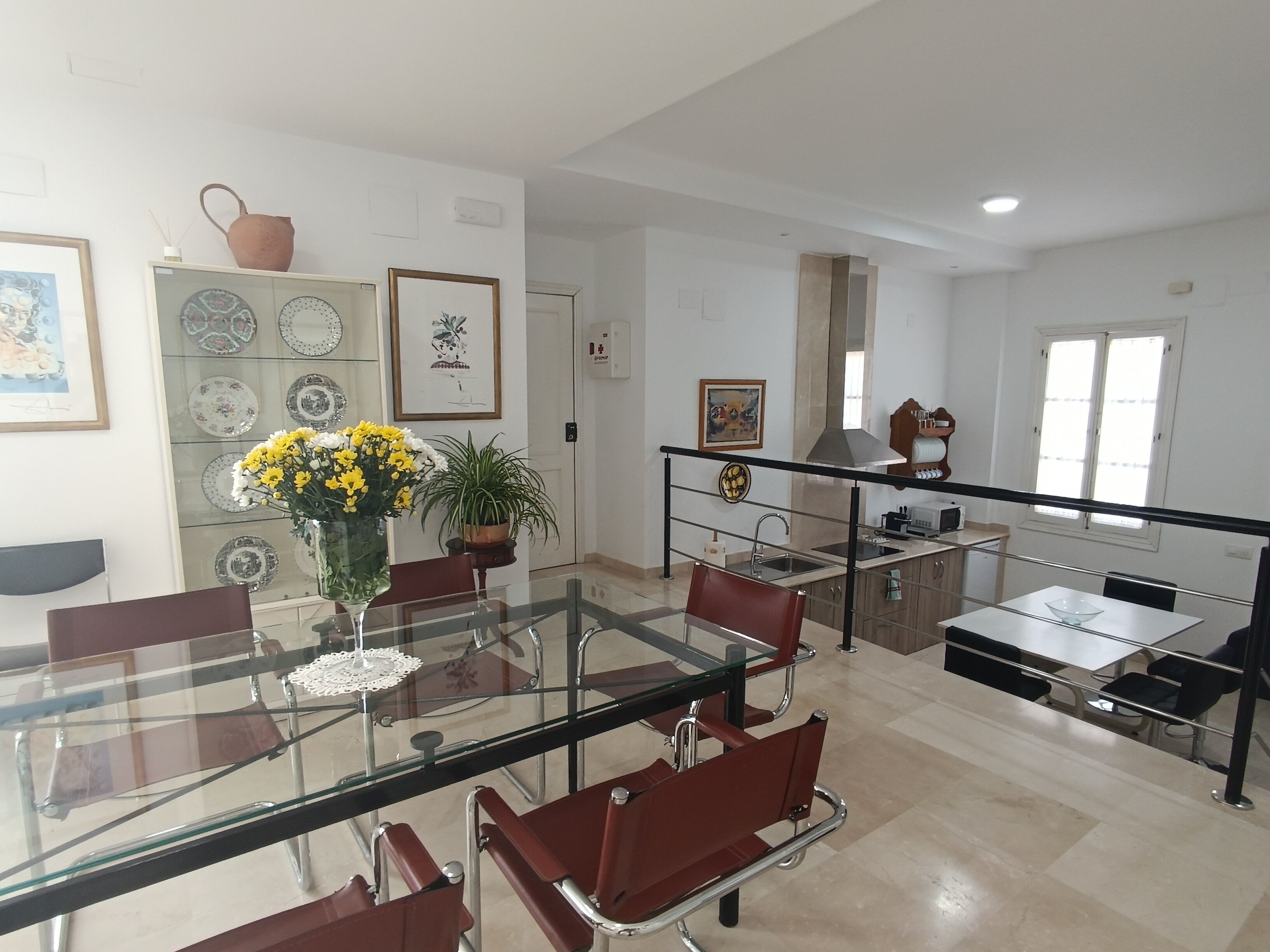A modern dining area with a glass-top table, red chairs, and a potted plant with yellow flowers.