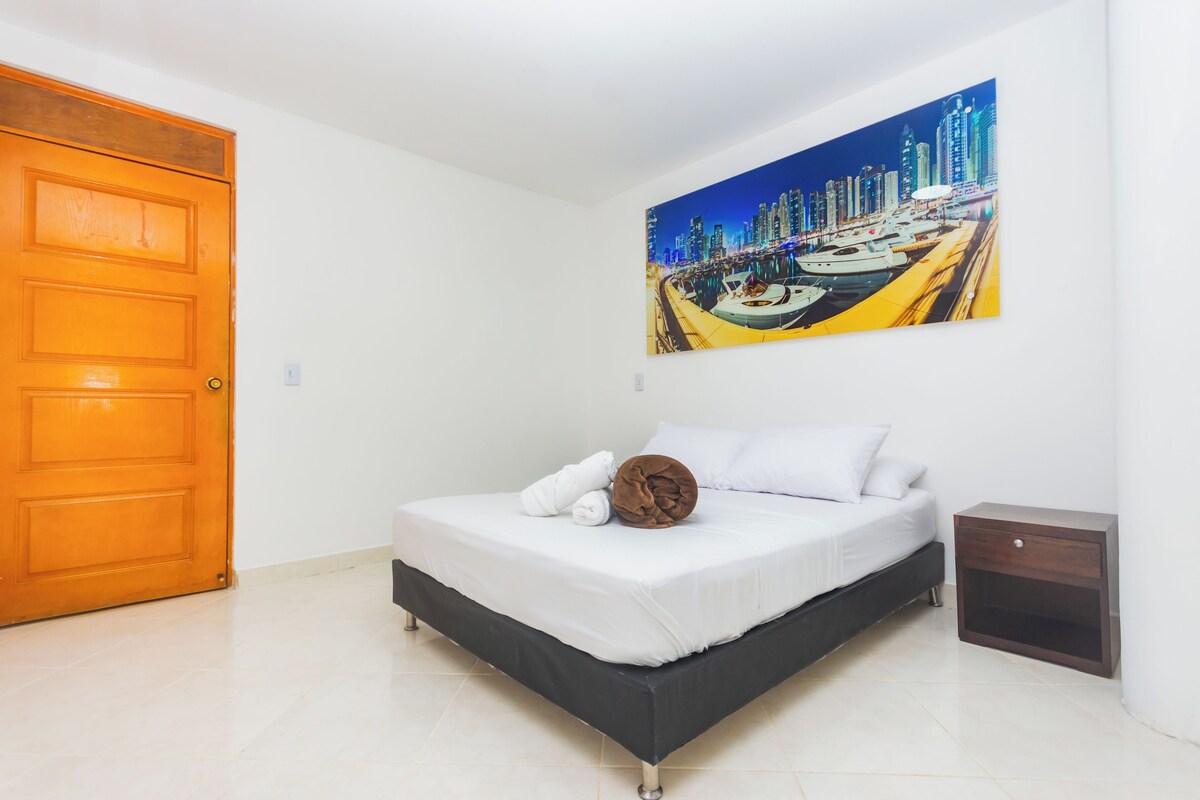 A bedroom with a bed, a nightstand, a painting of a cityscape, and a wooden door.