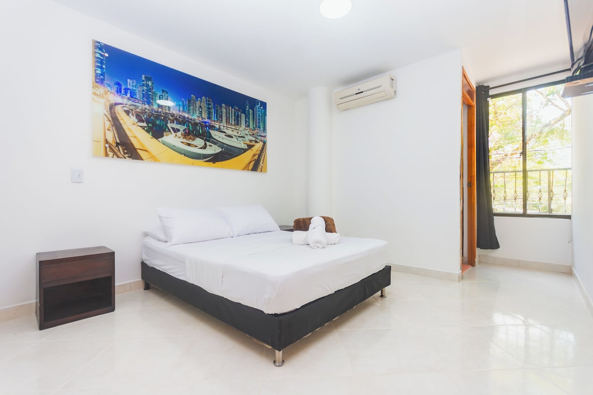 A hotel room with a bed, a nightstand, an air conditioning unit, and a large window with curtains.