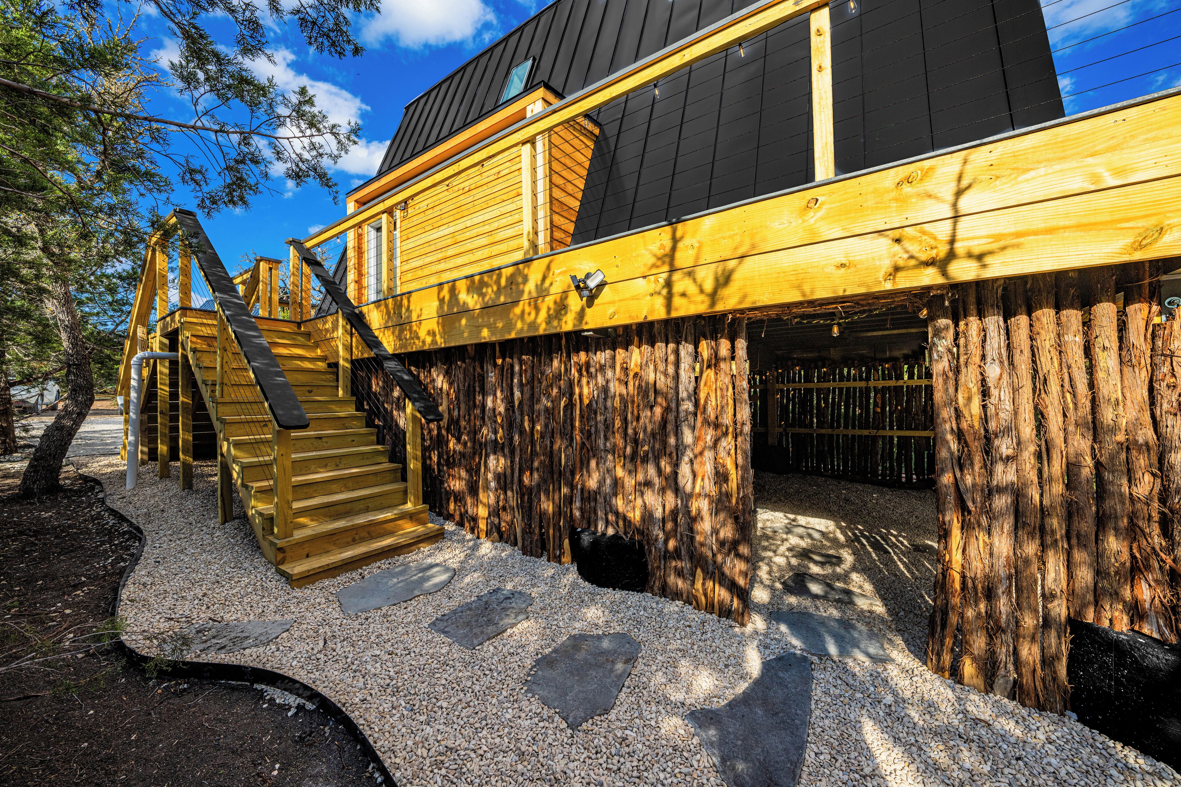 A wooden cabin with a staircase leading to a deck, surrounded by trees and gravel.