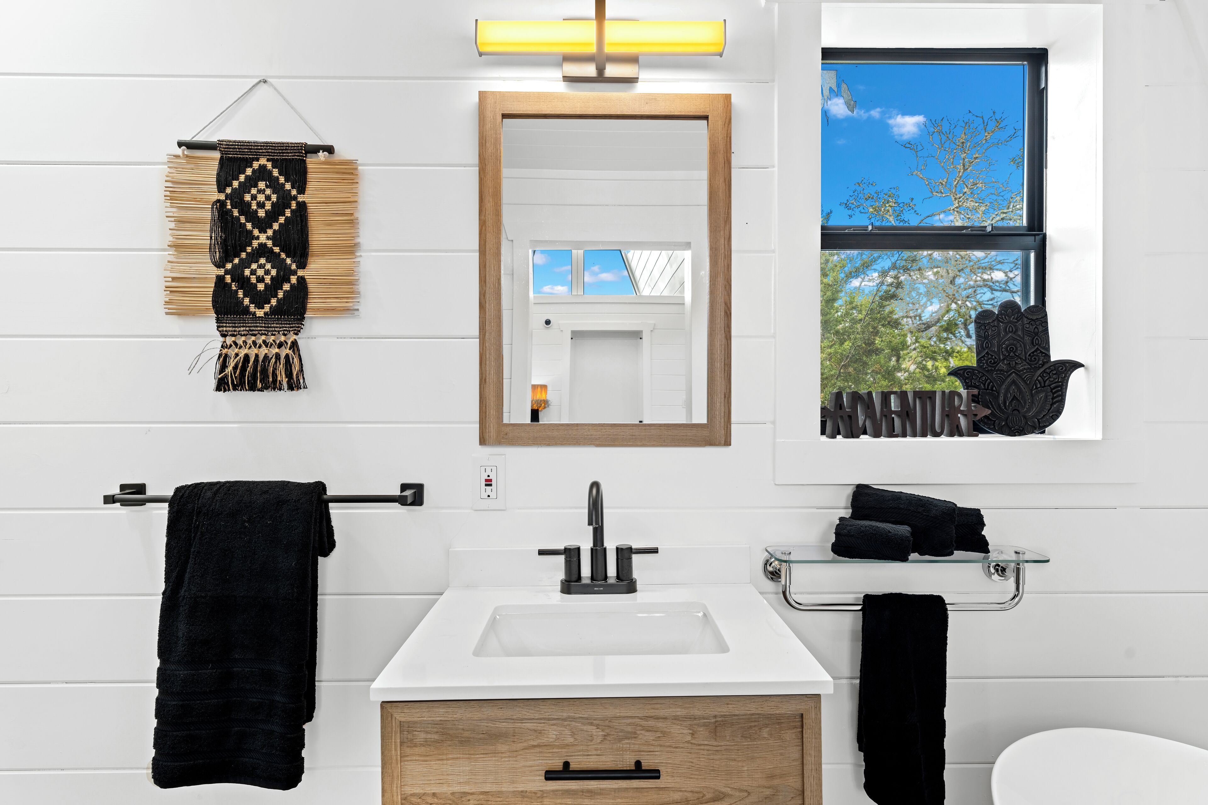 A modern bathroom with a white sink, black towel, wooden cabinet, and a window with a view of trees.