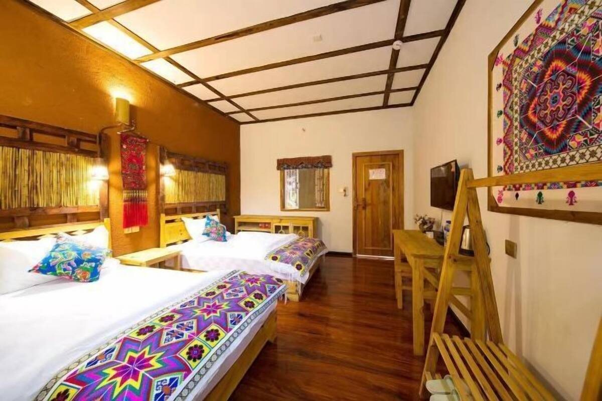 Quarto com duas camas de madeira, um banco de madeira, TV fixada na parede e uma tapeçaria colorida.