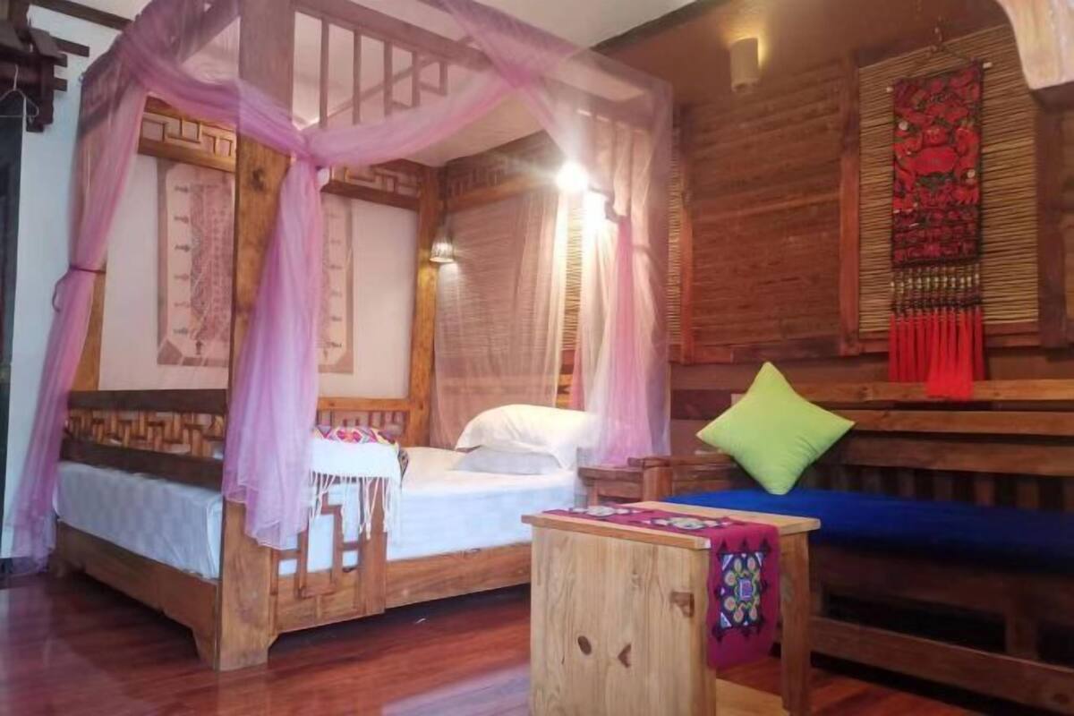 Uma cama de madeira com dossel e cortina, uma mesinha de madeira e um banco com almofadas.