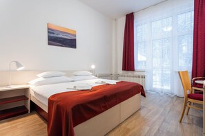 Habitación de hotel ordenada con cama, una silla, una mesa, una lámpara y un ventanal con cortinas.