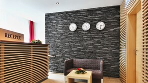 A reception area with a stone wall, three clocks, a reception desk, a brown sofa, and a small table with a flower arrangement. - Apartmánový Komplex Dagmar (Jachymov)