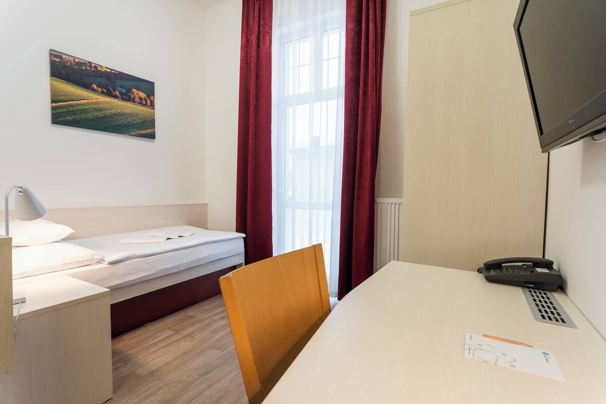 Une chambre d’hôtel avec un lit, un bureau, une chaise, une télévision et une fenêtre avec des rideaux.