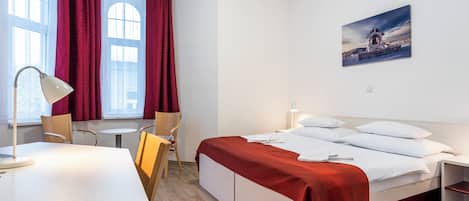 A hotel room with a bed, a desk, a chair, a small table, and a painting on the wall.