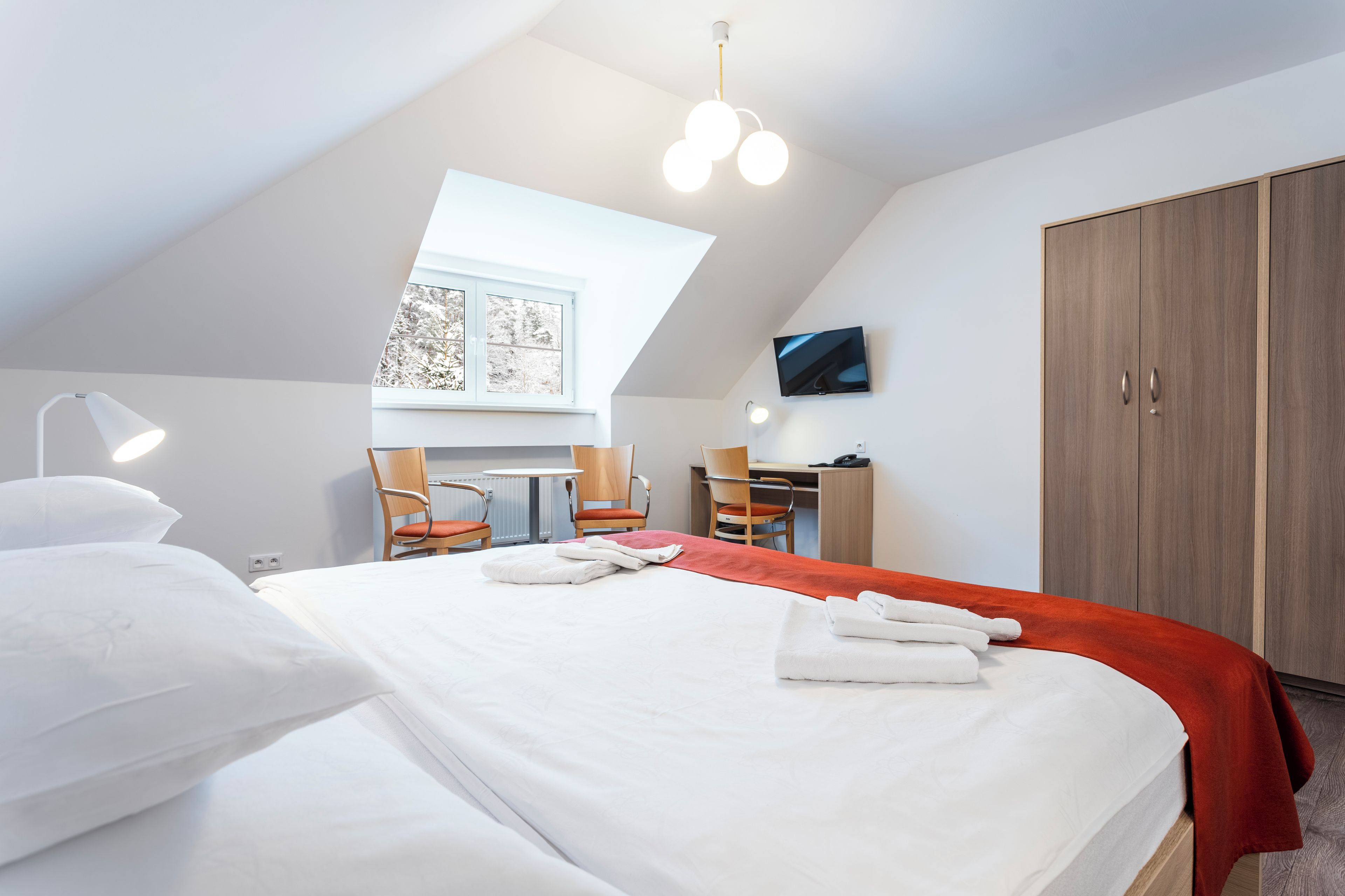 A hotel room with a large bed, a desk, two chairs, a TV, and a wardrobe.