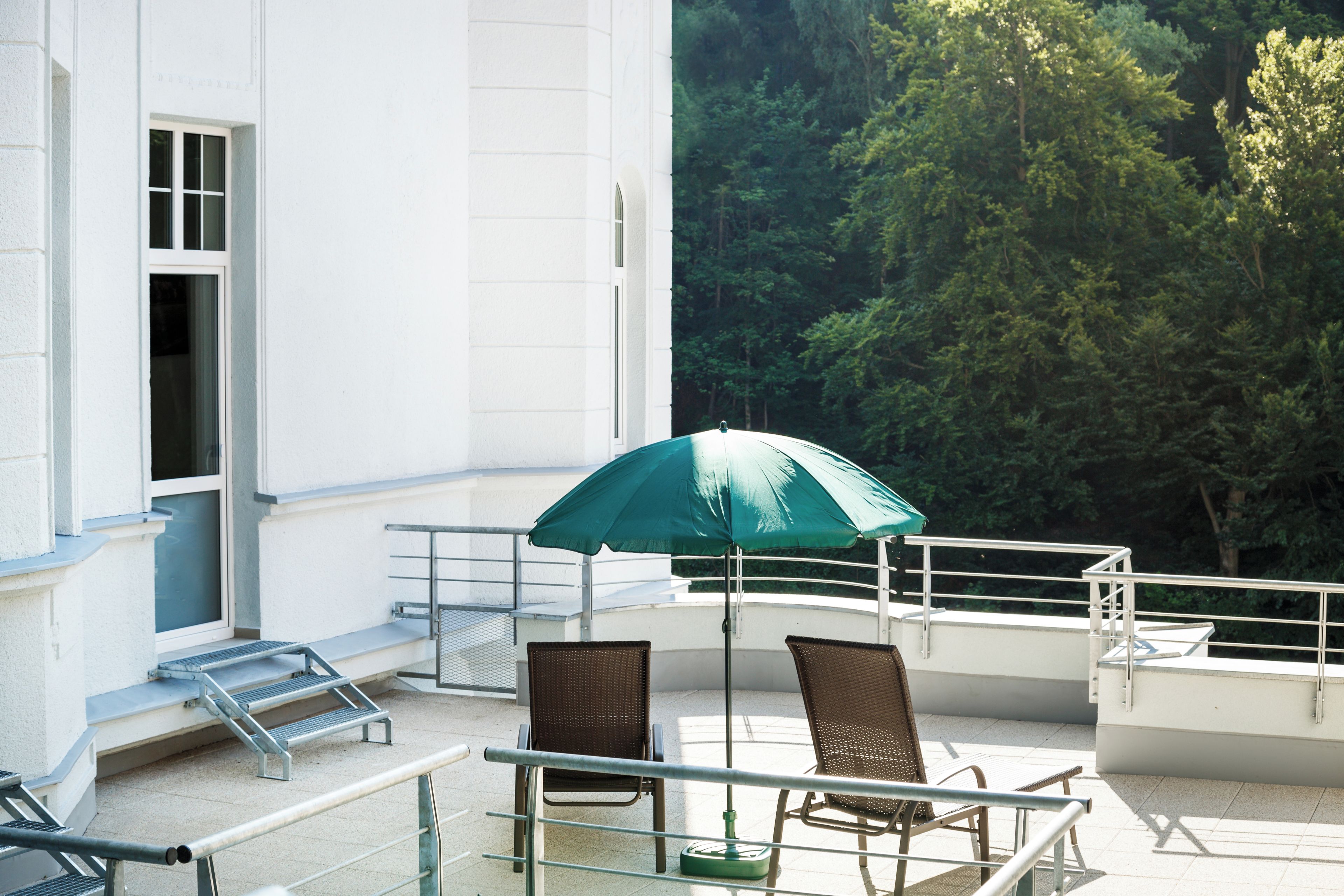 Ein Balkon mit einem grünen Sonnenschirm, zwei Korbstühlen und einem Glastisch.