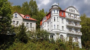 A large, white building with multiple balconies and a red roof, surrounded by lush greenery. - Apartmánový Komplex Dagmar (Jachymov)