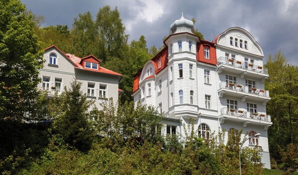 Un edificio grande y blanco con varios balcones y un techo rojo, rodeado de vegetación exuberante.