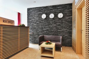 A reception area with a stone wall, three clocks, a reception desk, a brown sofa, and a small table with a flower arrangement. - Apartmánový Komplex Dagmar (Jachymov)