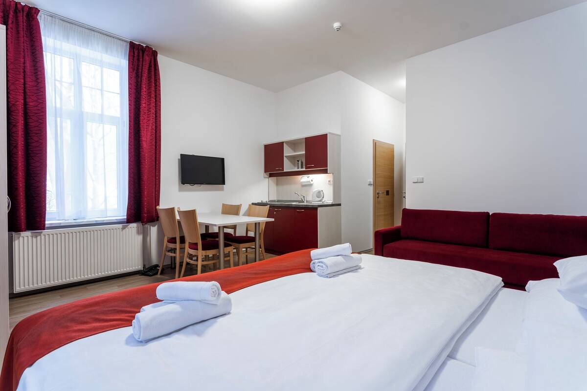 Une chambre d’hôtel avec un lit, un canapé rouge, un bureau avec une chaise, une petite kitchenette et un téléviseur fixé au mur.