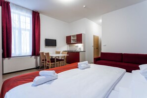 Habitación de hotel con cama, un sofá rojo, escritorio con silla, una kitchenette pequeña y un televisor en la pared.