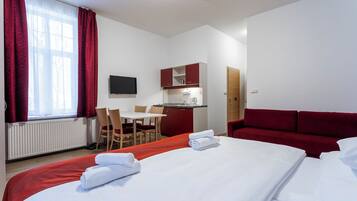 A hotel room with a bed, a red sofa, a desk with a chair, a small kitchenette, and a TV on the wall.