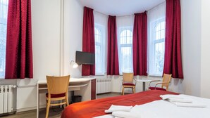A hotel room with a bed, two chairs, a desk, a TV, and large windows with red curtains. - Apartmánový Komplex Dagmar (Jachymov)