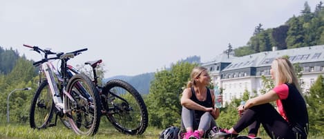 Two people sitting on grass near a mountain bike, with a large building in the background.
