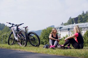 Two people sitting on grass near a mountain bike, with a large building in the background. - Apartmánový Komplex Dagmar (Jachymov)