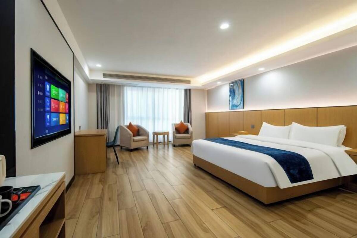 A modern hotel room with a large bed, a desk with a computer, and a TV displaying colorful icons.