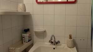Un baño con lavamanos blanco, espejo de marco rojo, inodoro y estante con artículos de aseo.