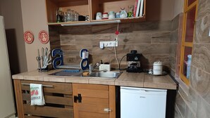 Una cocina con un lavamanos, cafetera y un refrigerador pequeño. Hay armarios de madera y un estante sobre la encimera.