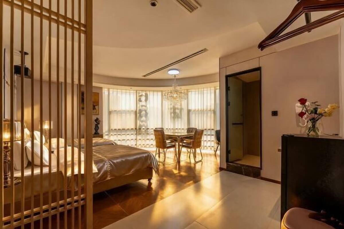 A bedroom with a bed, a desk, and a chair, a chandelier, and a view through a window.