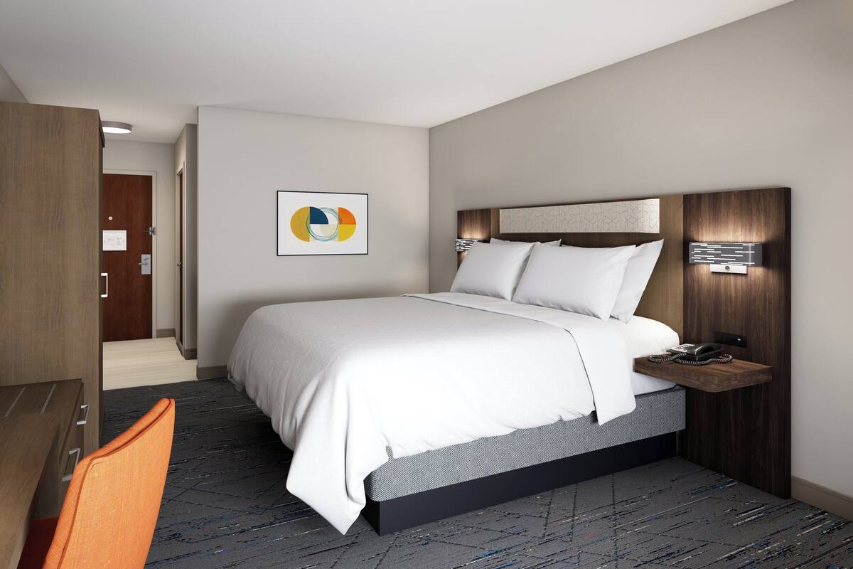 Ein modernes Hotelzimmer mit einem großen Bett, einem Schreibtisch und einem Sessel. Ein Nachttisch mit Telefon und Lampe steht bereit. Der Boden ist mit Teppich ausgelegt und an der Wand hängt ein Bild.
