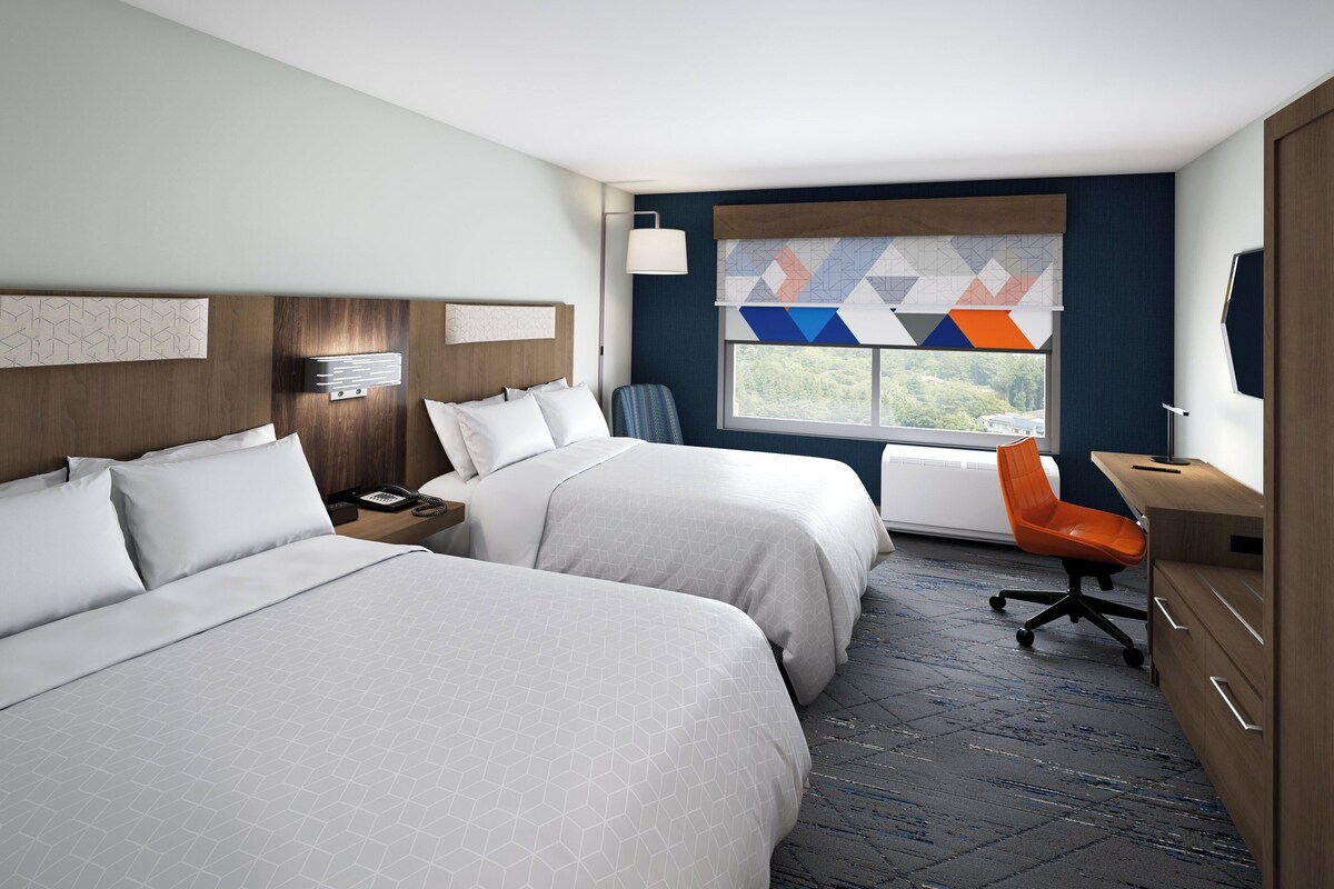 A hotel room with two beds, a desk, an orange chair, a window with a view, and a wall with geometric patterned curtains.