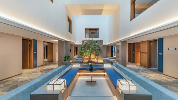 A spacious hotel lobby with a central water feature, multiple doors, and a large tree providing natural light.