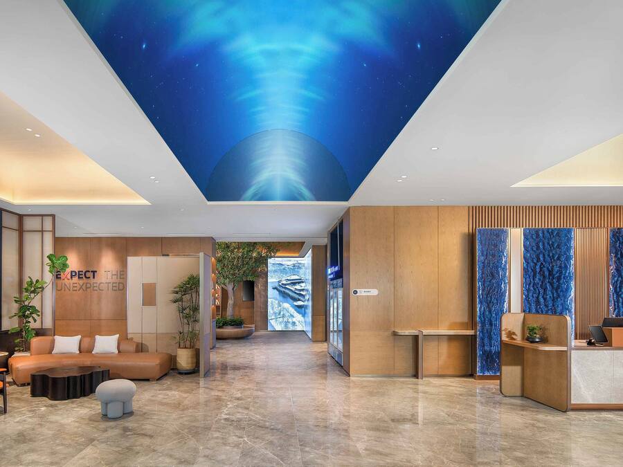 Un moderno vestíbulo de hotel con techo alto, una recepción, zona de estar y una pared con el letrero 'EXPECT THE UNEXPECTED'.