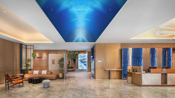 A modern hotel lobby with a high ceiling, a reception desk, seating area, and a wall with a 'EXPECT THE UNEXPECTED' sign.