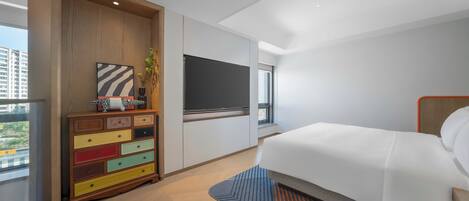Habitación de hotel moderna con cama, televisor, cómoda de colores y ventana con vista a edificios.