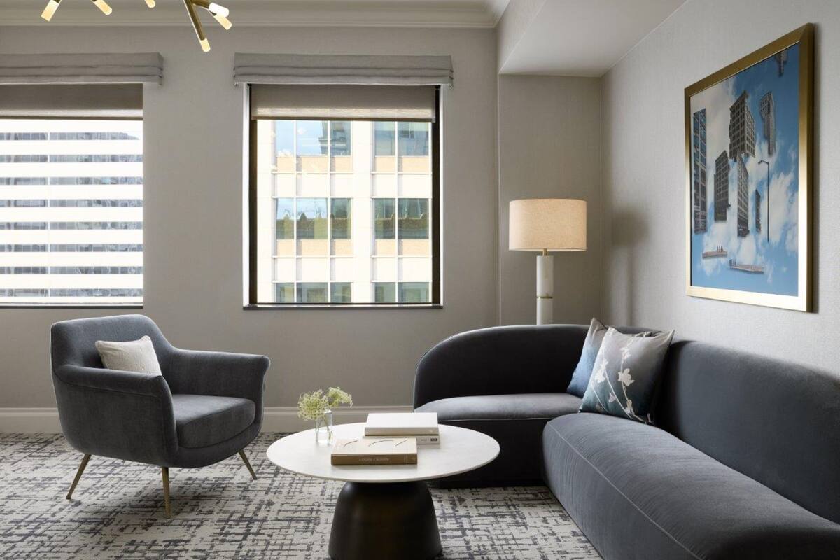 Una sala de estar moderna con un sofá y un sillón de color gris, y una mesa redonda. Hay una ventana con persianas, una lámpara de pie y un cuadro enmarcado en la pared.