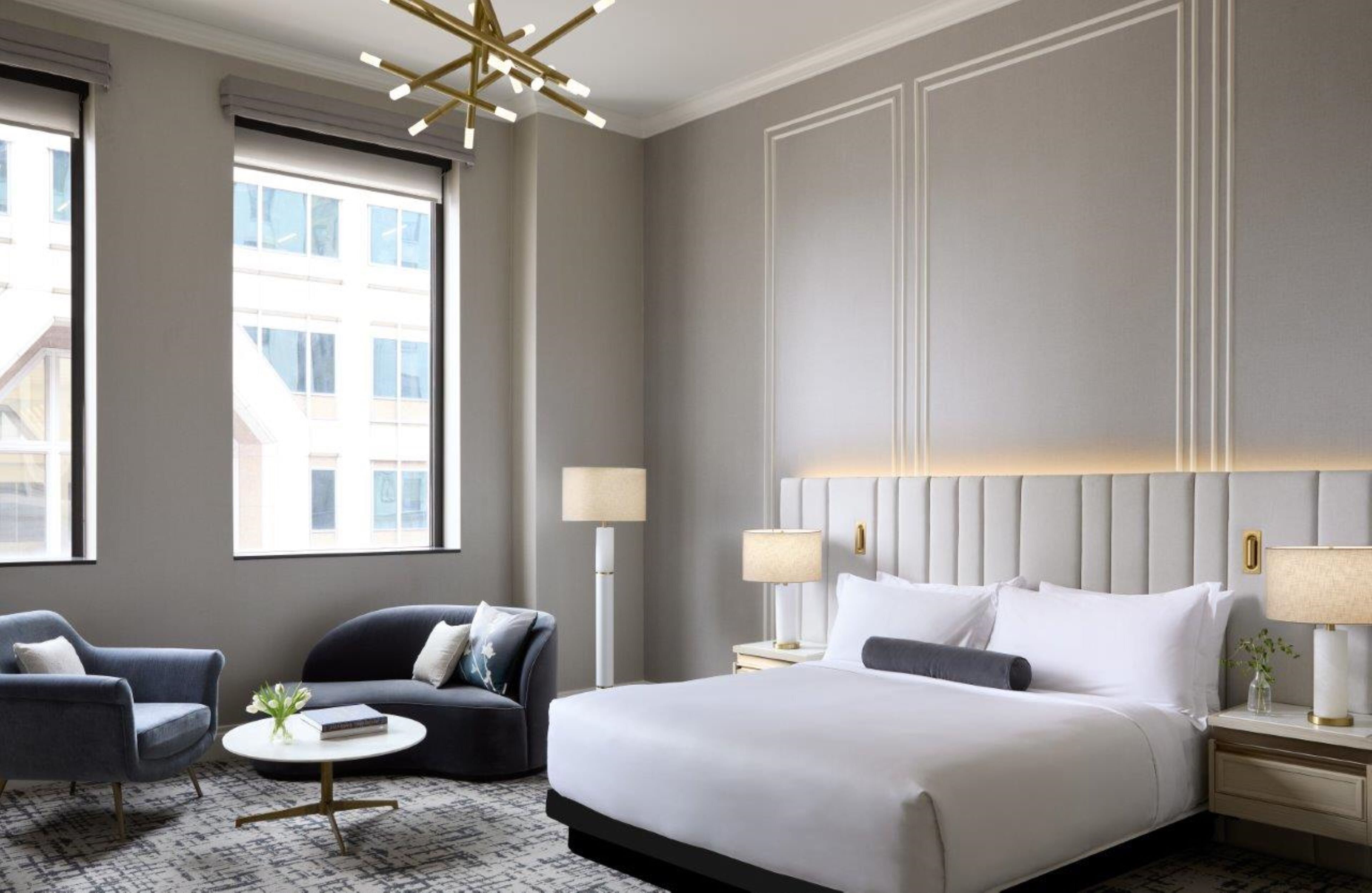 Een moderne hotelkamer met een groot bed, twee fauteuils, een salontafel en uitzicht op gebouwen door grote ramen.
