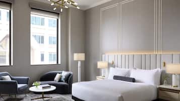 Een moderne hotelkamer met een groot bed, twee fauteuils, een salontafel en uitzicht op gebouwen door grote ramen.