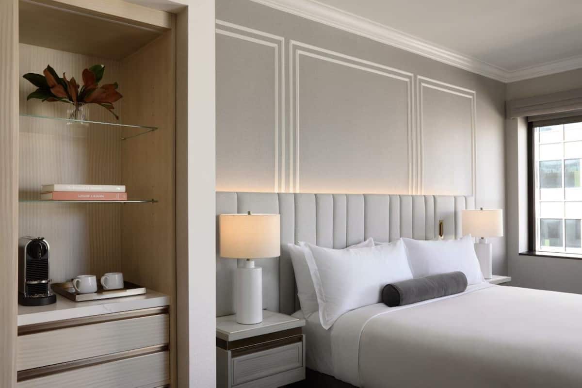 Habitación de hotel con cama, lámparas de noche, mesita de noche, estante con libros y cafetera, además de una ventana con vistas.