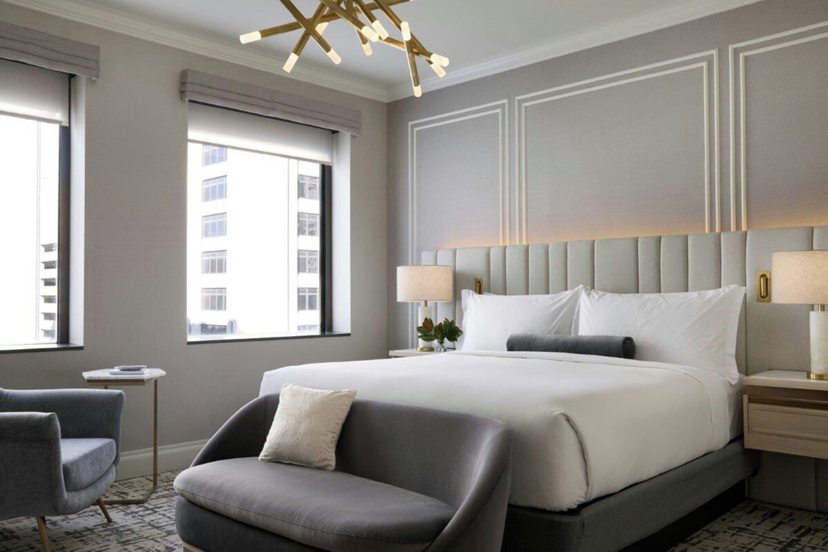 A modern hotel room with a large bed, a grey armchair, a bedside table with a lamp, and a view of the city through the window.